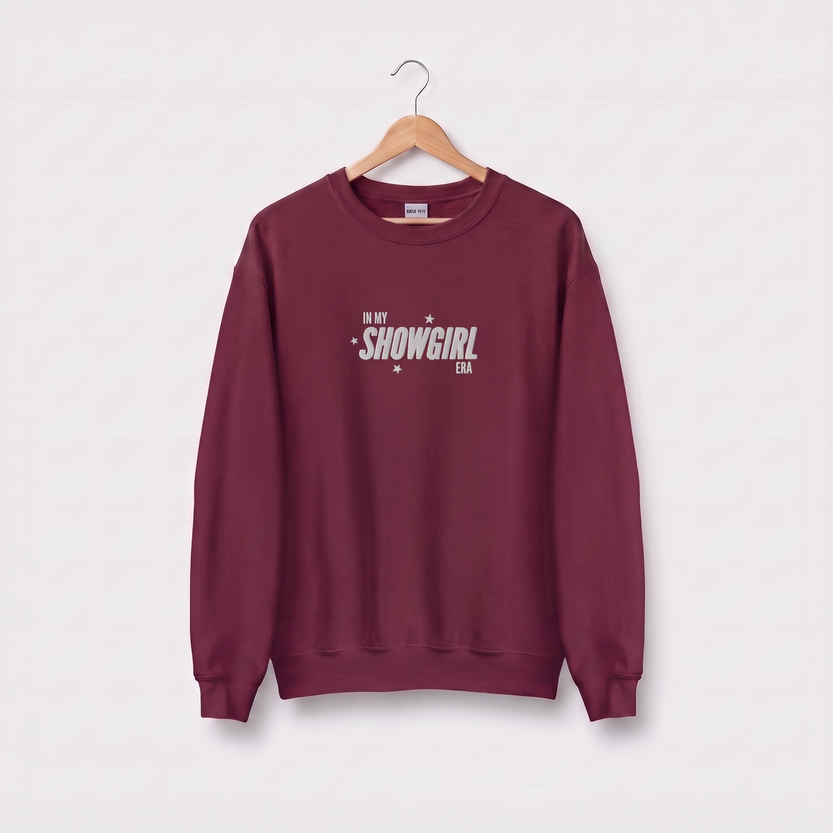A maroon crewneck sweatshirt hanging on a wooden hanger against a light gray background. The sweatshirt features the text 'IN MY SHOWGIRL ERA' printed in white on the chest.