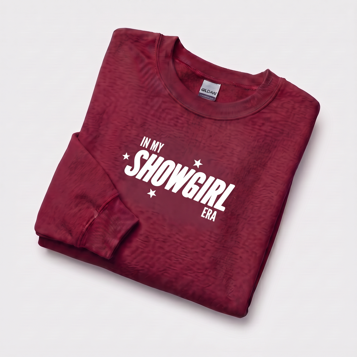 A folded maroon crewneck sweatshirt with a white graphic text print reading 'IN MY SHOWGIRL ERA' and small white stars, displayed on a light gray background. The neck label shows Gildan.