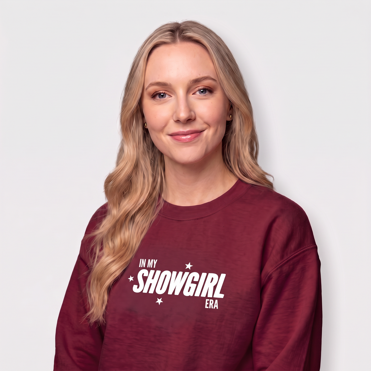 A woman with blonde hair wearing a maroon crewneck sweatshirt with white text that reads 'IN MY SHOWGIRL ERA' and small white stars, photographed against a light gray background.