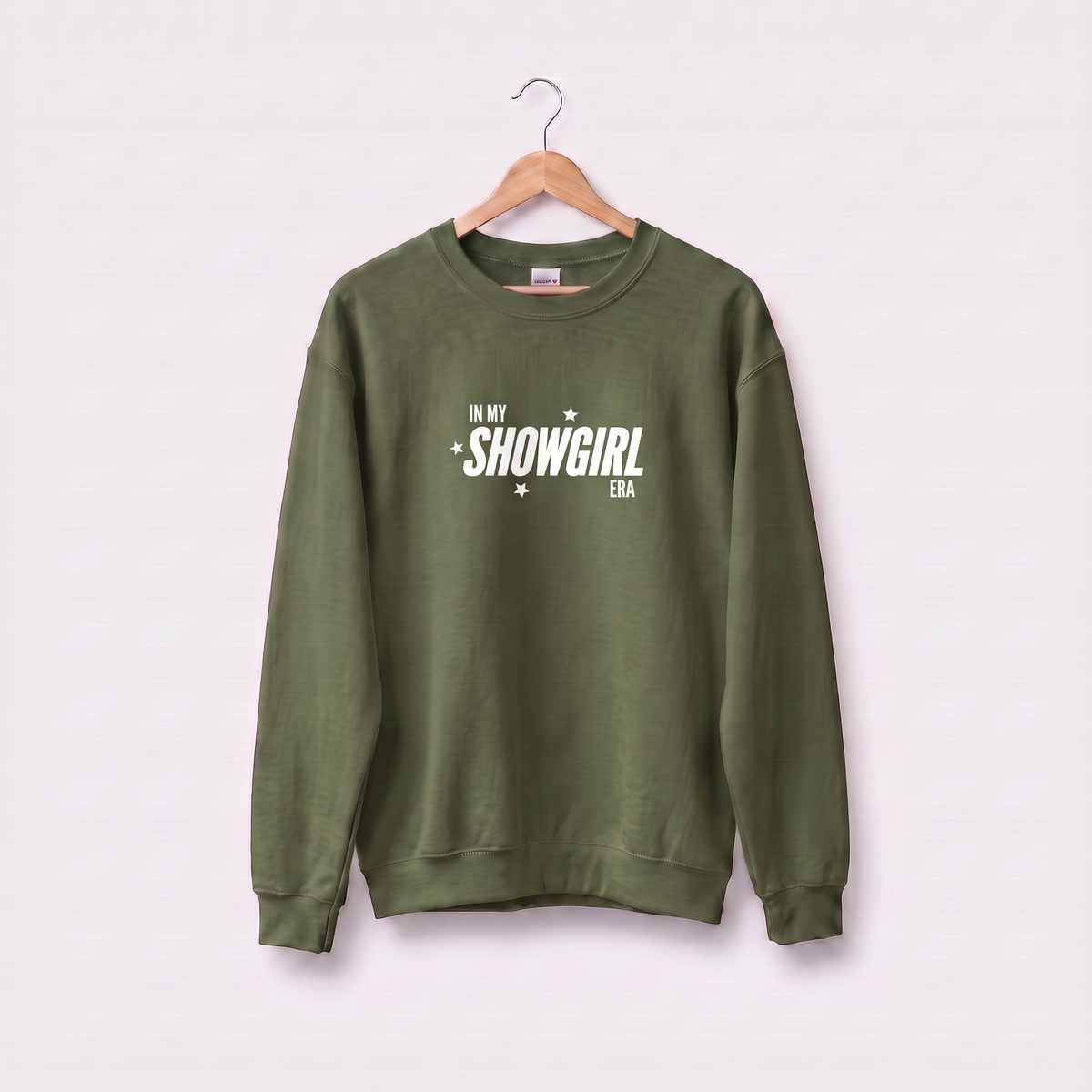 An olive green crewneck sweatshirt hanging on a wooden hanger against a light pink background. The sweatshirt features the text 'IN MY SHOWGIRL ERA' printed in white across the chest.