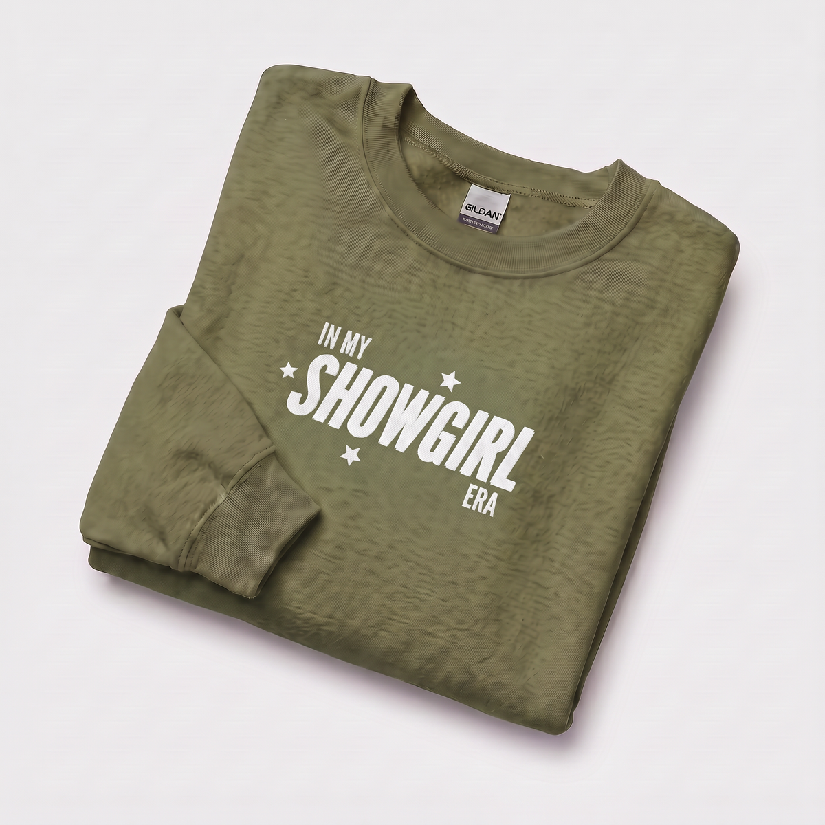 An olive green crewneck sweatshirt with a white graphic print reading 'IN MY SHOWGIRL ERA' and small white stars, folded neatly on a light gray background.