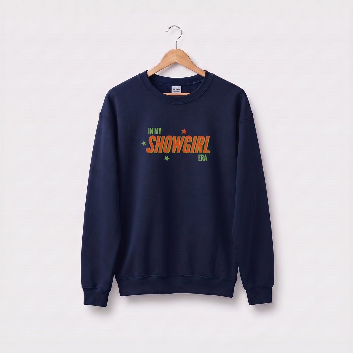 A navy blue crewneck sweatshirt hanging on a wooden hanger against a light gray background. The sweatshirt features a colorful graphic on the chest reading 'IN MY SHOWGIRL ERA' in orange and green lettering with small star accents.
