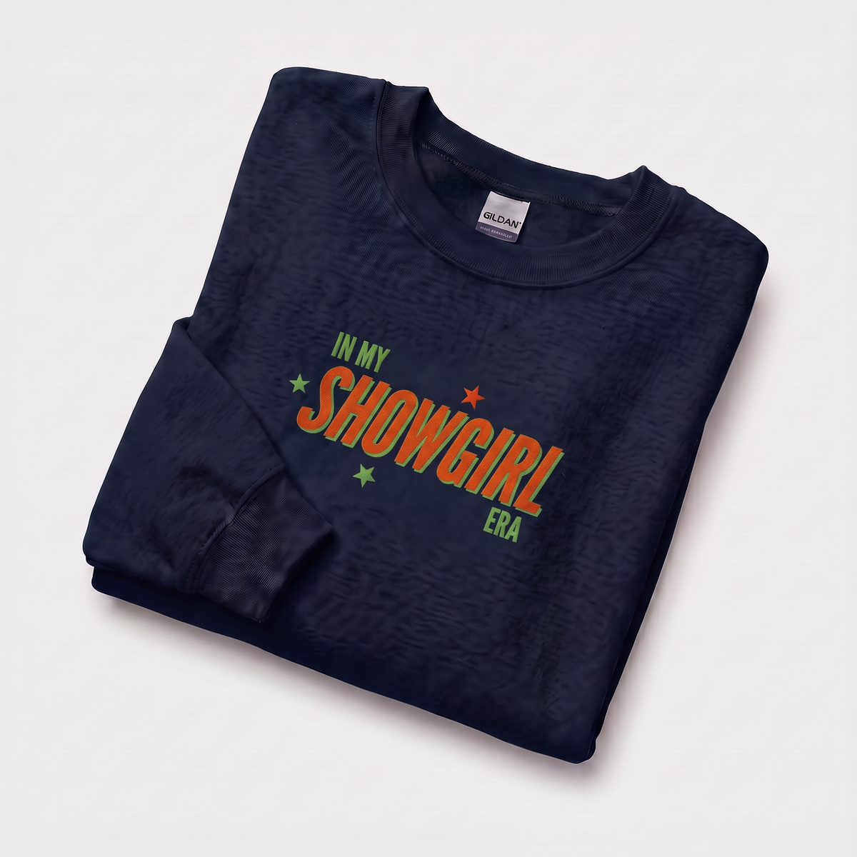 A folded navy blue crewneck with a colorful graphic reading 'IN MY SHOWGIRL ERA' in orange, green, and yellow, displayed on a clean white background.