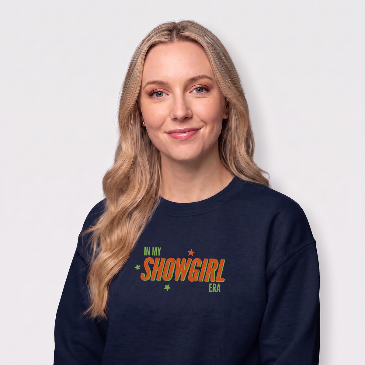 A woman with long blonde hair wearing a navy blue crewneck sweatshirt with a colorful graphic that reads 'IN MY SHOWGIRL ERA' in orange, green, and white, set against a plain light gray background.