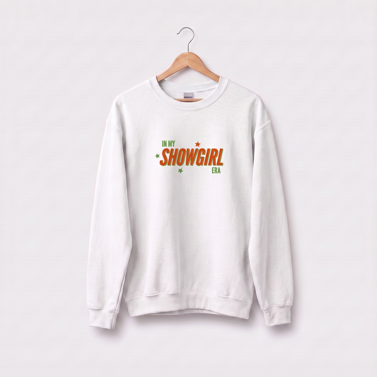 A white crewneck sweatshirt hanging on a wooden hanger against a light gray background. The sweatshirt features a graphic on the chest that reads 'IN MY SHOWGIRL ERA' in orange and green lettering.