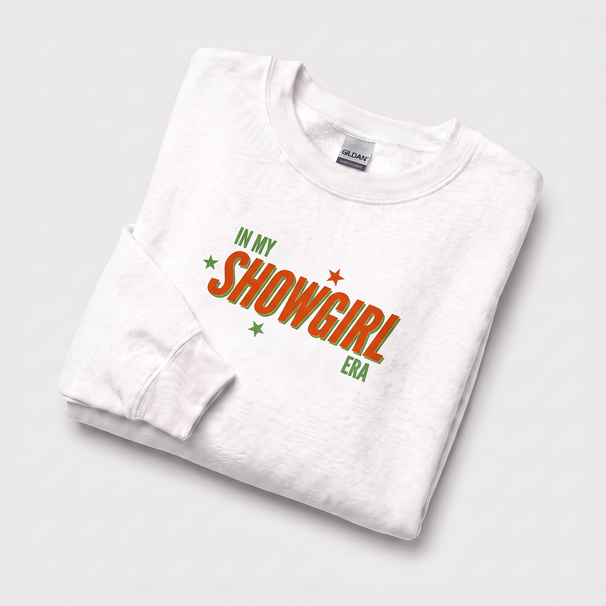 A folded white long-sleeve crewneck sweatshirt on a light gray background, featuring a graphic print that reads 'IN MY SHOWGIRL ERA' in green and orange lettering with small star accents.
