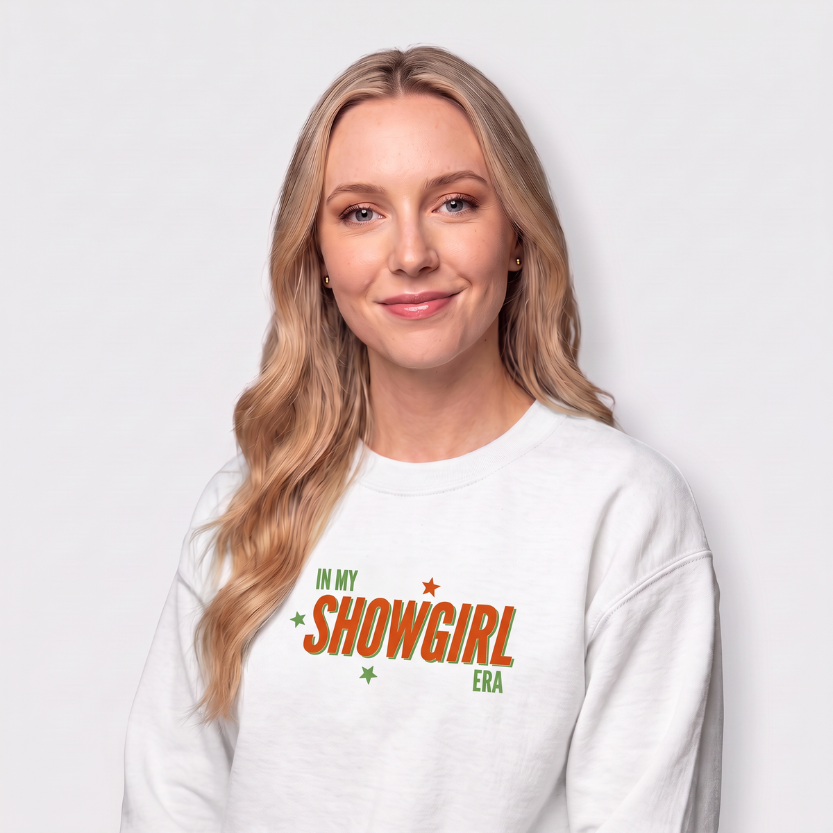 A blonde woman smiling while wearing a white crewneck sweatshirt with 'IN MY SHOWGIRL ERA' printed in gold text on the chest, set against a plain light gray background.