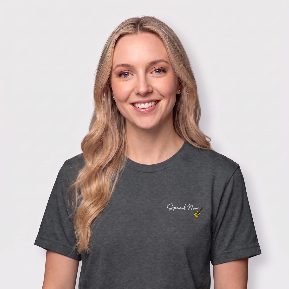 A smiling blonde woman wearing a dark heather gray t-shirt with a small white 'Speak Now' logo and a tiny yellow guitar graphic on the left chest, set against a plain light gray background.