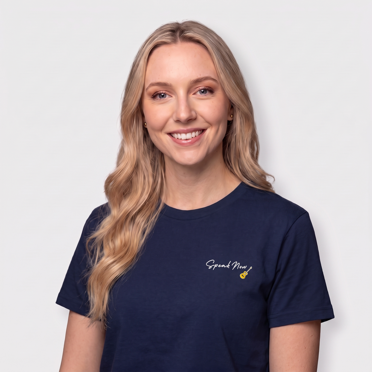 A smiling blonde woman wearing a navy blue crewneck t-shirt with the text 'Speak Now' printed in white on the left chest, set against a plain light gray background.