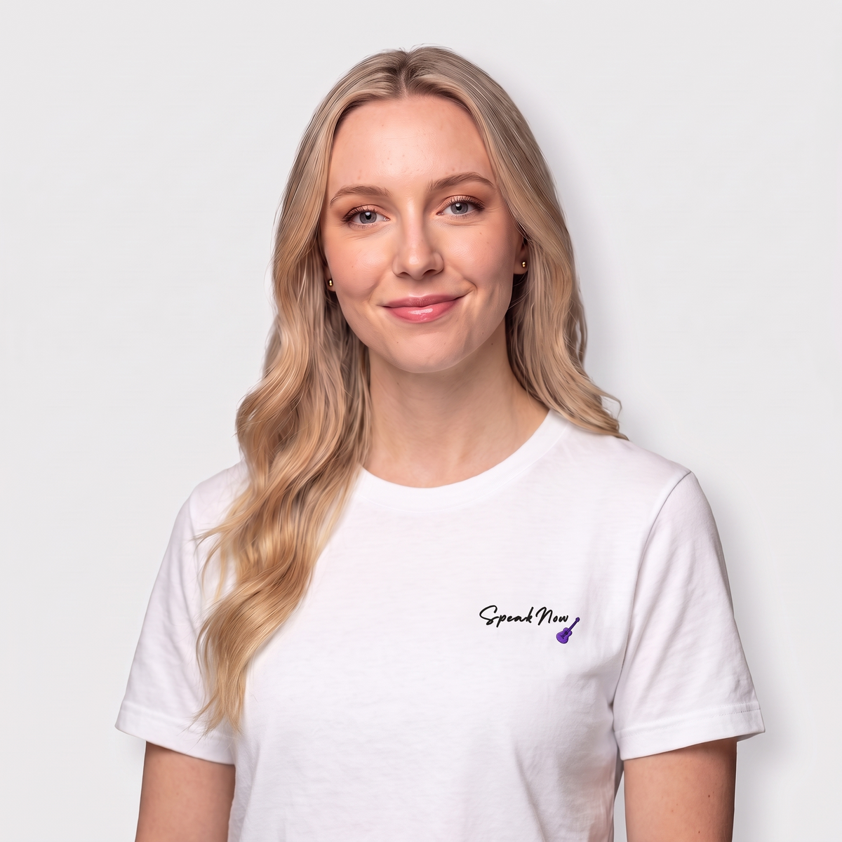 A blonde woman with blue eyes wearing a white crewneck t-shirt with a small black 'Spread Now' logo and a purple heart on the left chest, set against a plain light gray background.
