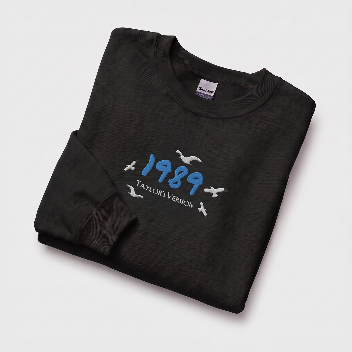 A folded black long-sleeve t-shirt with a graphic print on the front. The graphic features the year '1989' in blue, 'TAYLOR'S VERSION' in white, and several white seagull silhouettes.