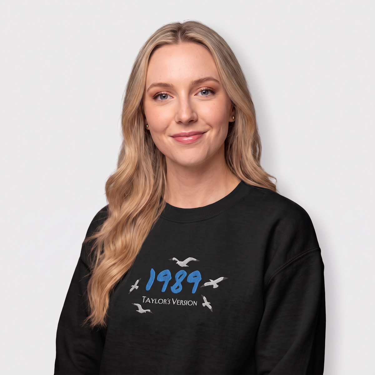 A blonde woman wearing a black crewneck sweatshirt with a graphic on the chest that reads '1989 TAYLOR'S VERSION' with white seagull silhouettes, set against a plain light gray background.