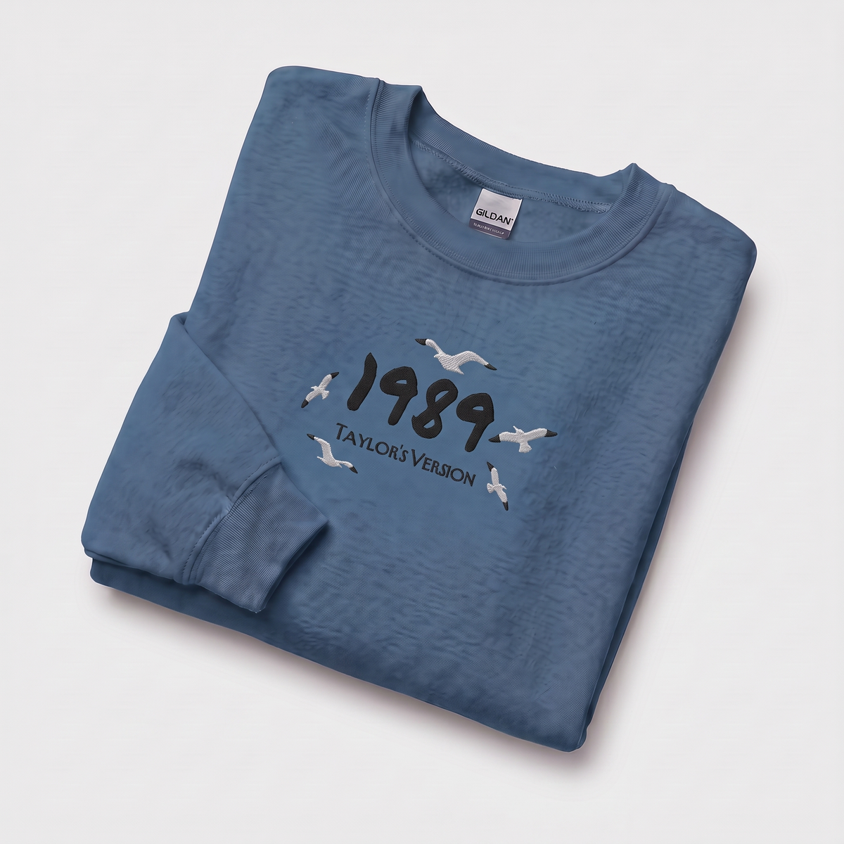 A folded blue long-sleeve t-shirt with a graphic on the front reading '1989 TAYLOR'S VERSION' with seagull illustrations, displayed on a light gray background. The neck label shows the brand Gildan.