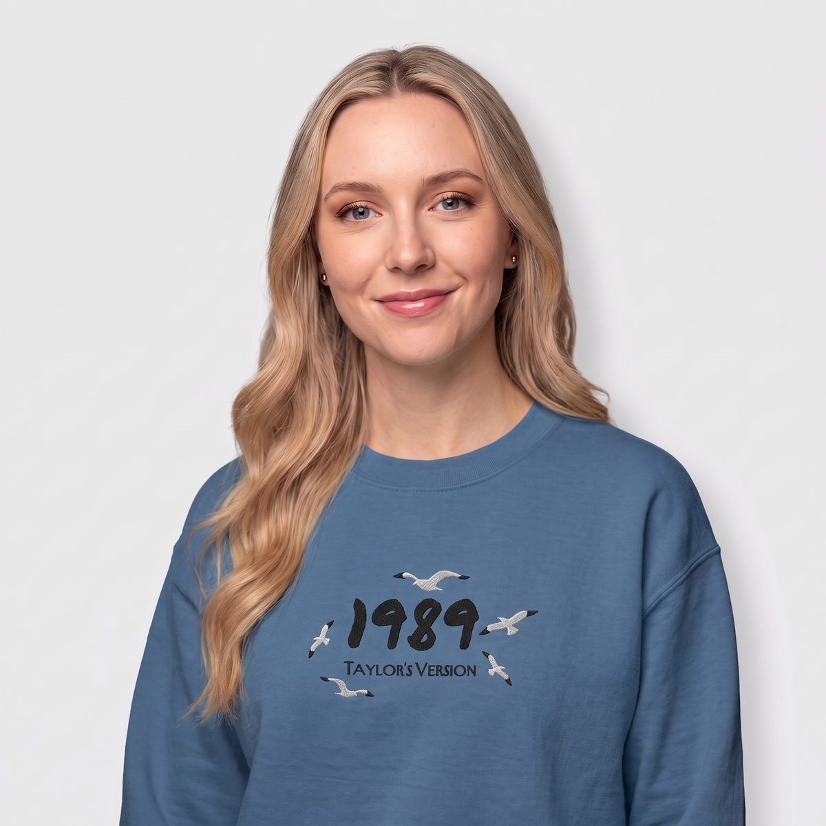 A woman with blonde hair wearing a slate blue crewneck sweatshirt with a graphic print of flying seagulls and the text '1989 TAYLORS VERSION'.