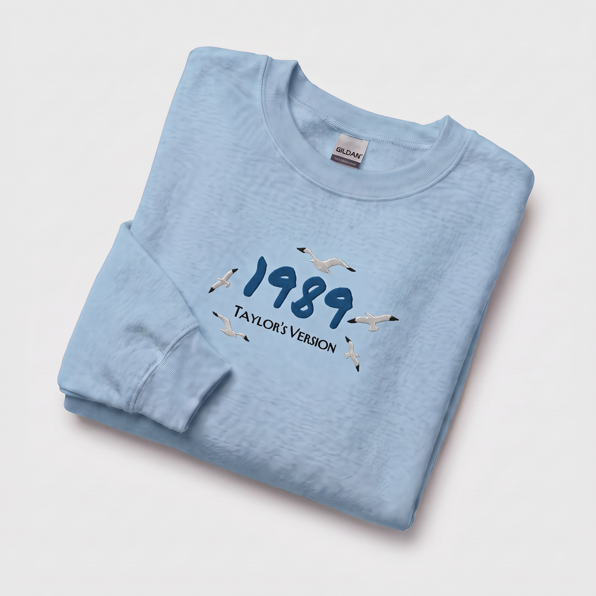 A folded light blue crewneck sweatshirt on a light gray background, featuring a graphic print of the year '1989' in navy blue with seagulls flying around it and the text 'TAYLOR'S VERSION'. The inside collar shows a visible Gildan brand tag.
