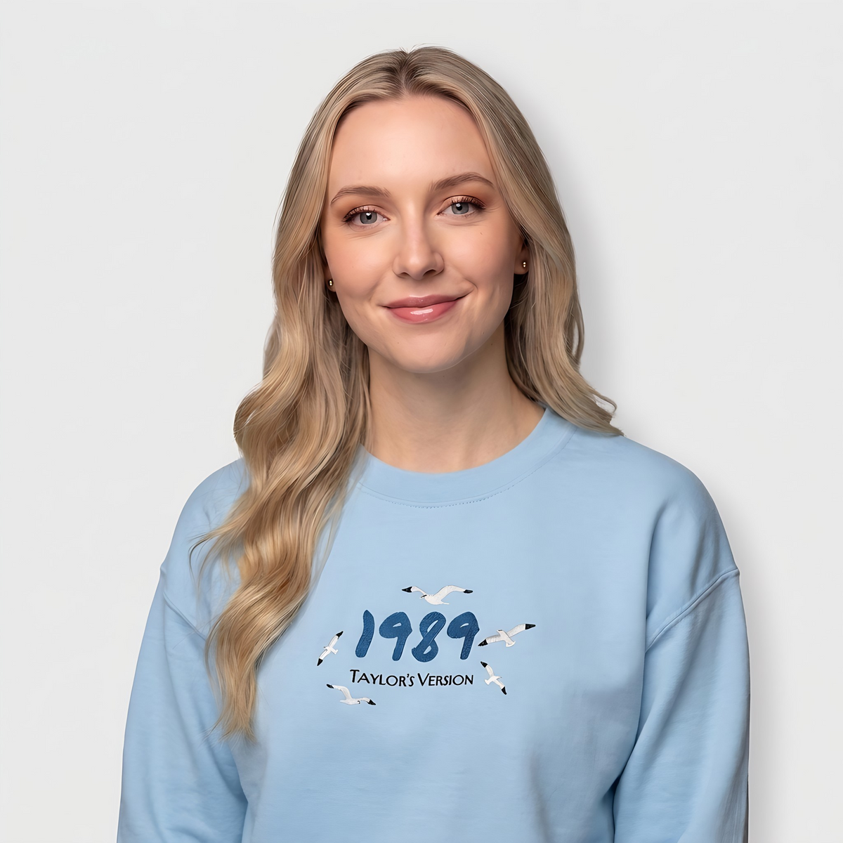 A blonde woman wearing a light blue crewneck sweatshirt with a graphic on the chest that reads '1989 TAYLOR VERSION' surrounded by small bird illustrations, set against a plain light gray background.