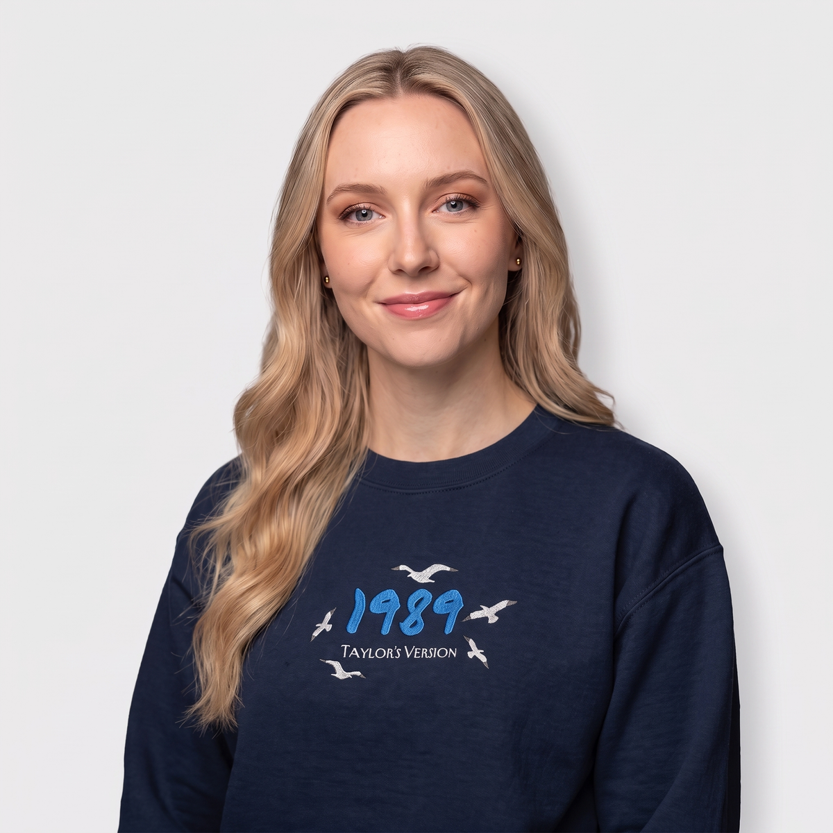 A blonde woman wearing a navy blue crewneck sweatshirt with a graphic on the chest that reads '1989 TAYLOR'S VERSION' with white seagull silhouettes, set against a plain light gray background.